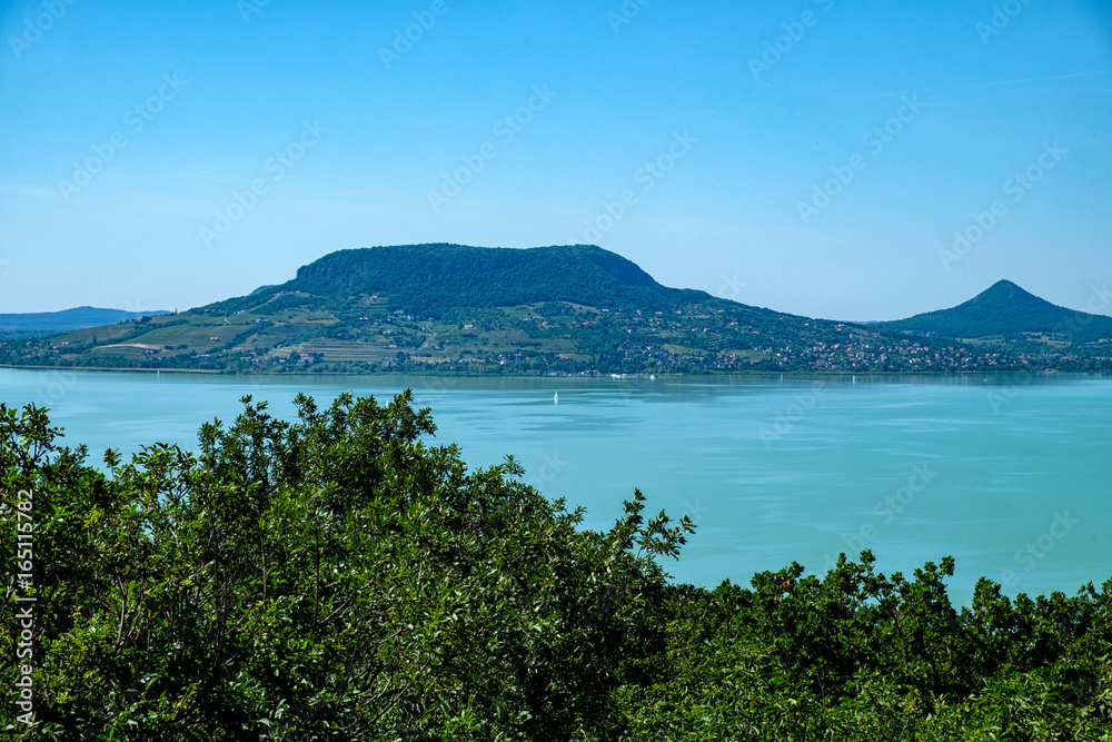 sailboat on water, greenery in foreground fameus Badacsony hill in background - summertime and vacation at Balaton lake