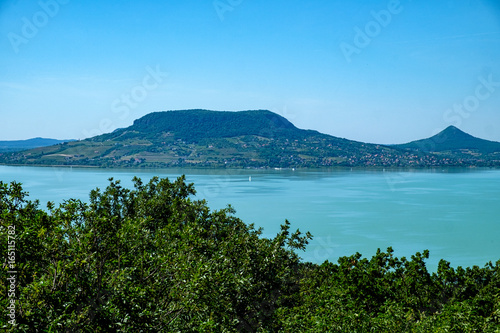 sailboat on water, greenery in foreground fameus Badacsony hill in background - summertime and vacation at Balaton lake