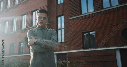 confident hipster man guy boy arms crossed crossing moving camera looking round sunglasses grey shirt light beam sunny evening building red cool fashionable hands watch clock rays lit ray balanced photo