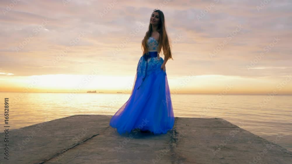 The young beautiful girl dances in a blue evening or ball dress on the pier close to sea. Slow motion. The young beautiful girl dances in a blue evening or ball dress on the pier close to sea. Slow motion.