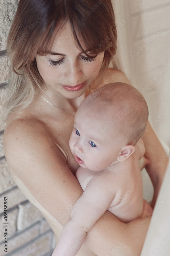 Young mother looking at her little blue-eyed baby wearing nothing relaxing and playing in the bedroom at the weekend together, lazy morning, warm and cozy scene. Brick wall background. Young mother looking at her little blue-eyed baby wearing nothing relaxing and playing in the bedroom at the weekend together, lazy morning, warm and cozy scene. Brick wall background.