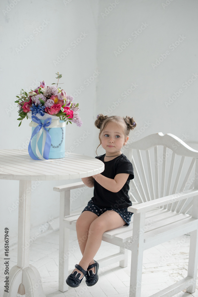 Cute baby at home in white room is sitting on a white chair. The beautiful girl with a beautiful box of flowers on the table. Baby with curly hair is looking away Cute baby at home in white room is sitting on a white chair. The beautiful girl with a beautiful box of flowers on the table. Baby with curly hair is looking away