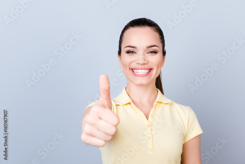 I like it! Young cute girl, standing on the pure light blue background and smiling, wearing a yellow tshirt and showing thumbup sign