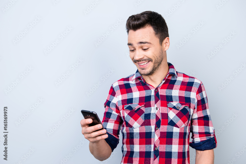 Young cheerful brunet student is standing on the pure light blue background and smiling amazed, reading good news on internet at his mobile phone Young cheerful brunet student is standing on the pure light blue background and smiling amazed, reading good news on internet at his mobile phone