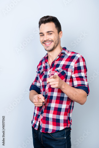 Smiling young stylish bearded brunet student in bright casual checkered shirt is standing on light background and point on camera with flirty smile and glance photo