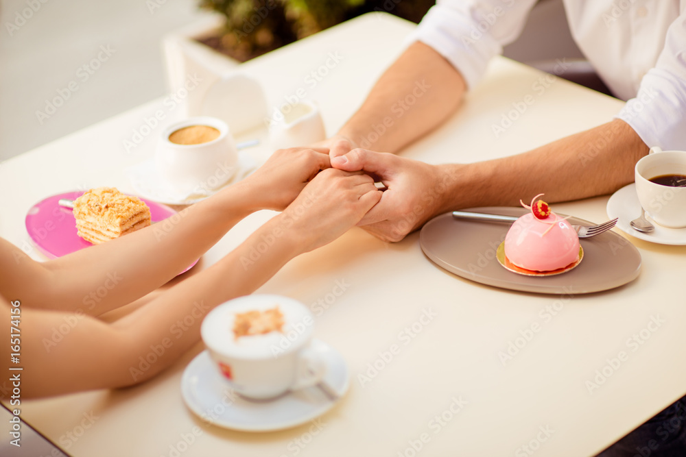 Close up cropped photo of two young lovers, sitting in cafe and hold each other`s hands, having nice coffees with delicious cakes