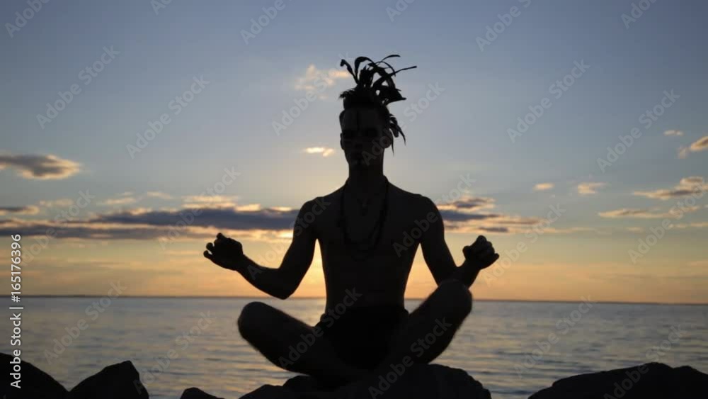 Silhouette of a man with native american indian feather mohawk accessory on head practicing yoga at the sunset.