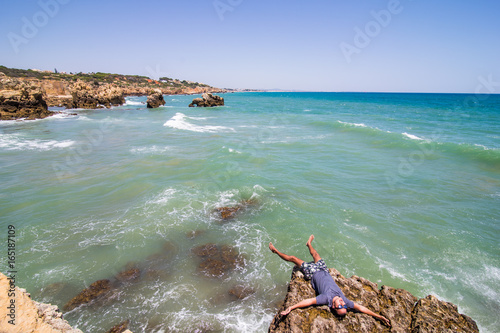 Young man lying on the rocks cliffs over the ocean of algarve portugal. summer vocation concept
