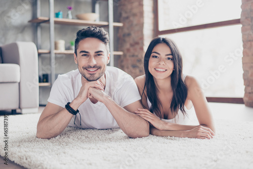 True love. Happy beautiful married latino couple is chilling out lying on the carpet at the floor at home indoors, looking at the camera and smiling
