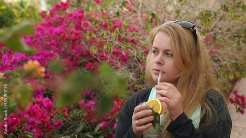 Young woman with long blond hair drinking a cold Mojito on a background of bright flowers in the Park of the hotel. Girl drinking cocktail from the straw in the Park. Slow motion photo