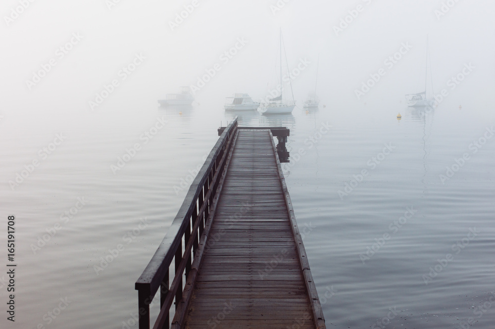 Wooden jetty stretching out onto a river with moored yachts just visible in dense fog Wooden jetty stretching out onto a river with moored yachts just visible in dense fog