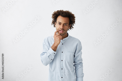 Indoor picture of serious pensive young black European male office worker rubbing face at mouth while thinking over working ideas and solutions, looking at camera, deep in thoughts. Body language