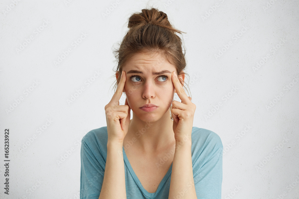 Attractive blue-eyed female with hair bun holding fingers on temples and looking aside trying to find solution of her trouble. Beautiful woman with dark hair trying to focuse on something important Attractive blue-eyed female with hair bun holding fingers on temples and looking aside trying to find solution of her trouble. Beautiful woman with dark hair trying to focuse on something important