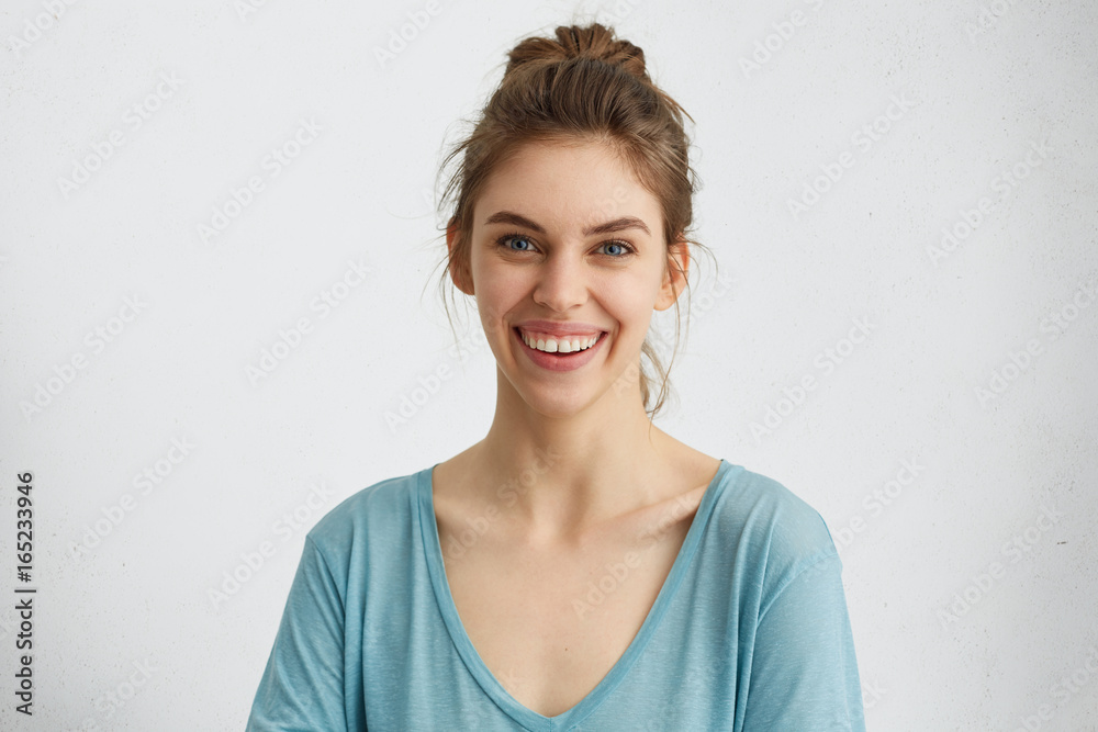 Happy smiling female with attractive appearance and dark hair tied in knot wearing blue blouse showing her broad smile having good mood liking to pose at camera. Happiness, positiveness concept Happy smiling female with attractive appearance and dark hair tied in knot wearing blue blouse showing her broad smile having good mood liking to pose at camera. Happiness, positiveness concept