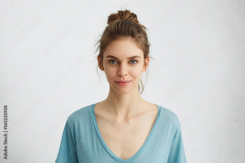 Portrait of beautiful female having blue shining eyes, thin lips, pure skin and dark hair tied in knot wearing blue loose blouse looking directly into camera having mysterious and glad expression