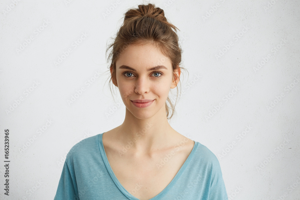 Horizontal portrait of attractive blue-eyed female dressed casually having delightful look while smiling. Beautiful woman with hair bun having cheerful expression while posing at studio over white Horizontal portrait of attractive blue-eyed female dressed casually having delightful look while smiling. Beautiful woman with hair bun having cheerful expression while posing at studio over white