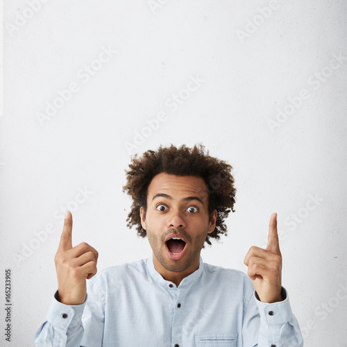 Vertical portrait of Afro American bug-eyed male being shocked from something, dressed in white shirt, pointing his finger at blank wall with copy space for your advertising content or hearder photo