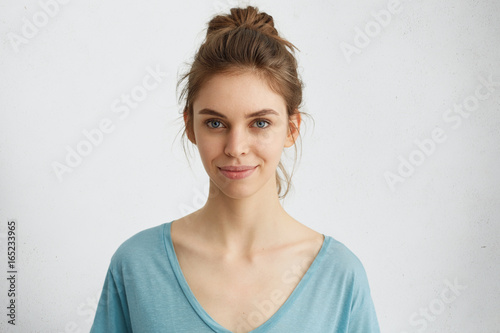 Horizontal portrait of attractive blue-eyed female dressed casually having delightful look while smiling. Beautiful woman with hair bun having cheerful expression while posing at studio over white