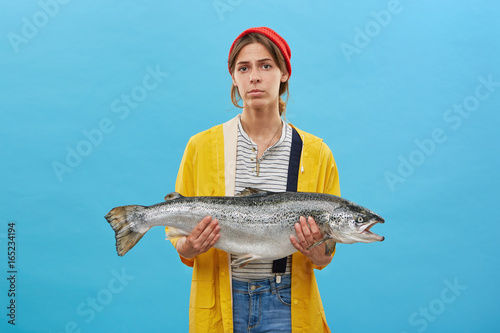 Young fisherwoman wearing red hat, yellow raincoat and jean overalls holding huge fish presenting her catch. Attractive woman buying big salmon or trout. Successful female angler posing with her catch