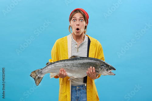 Portrait of shocked female with widely opened eyes and mouth holding huge fish which her husband caught isolated over blue background. Scary female angler with big catch. Fishing, recreation concept