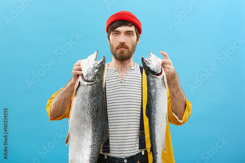 Confident man with beard, wearing red casual hat, sweater, overalls and yellow raincoat holding his big catch spending his free time at lake or river fishing all day long having great desire
