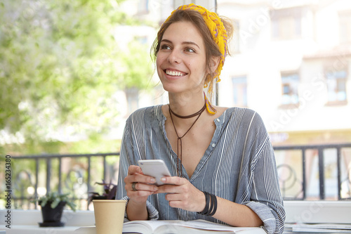 Attractive cheerful young Caucasian woman with charming smile wearing yellow headband sitting at terrace with book and coffee in papercup, reading text message on mobile phone during breakfast