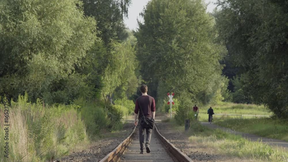 Walking Into An Uncertain Future. Young Man Walking Away From The Camera In Between Railroad Tracks In A Rural Sunny Summer Landscape Walking Into An Uncertain Future. Young Man Walking Away From The Camera In Between Railroad Tracks In A Rural Sunny Summer Landscape