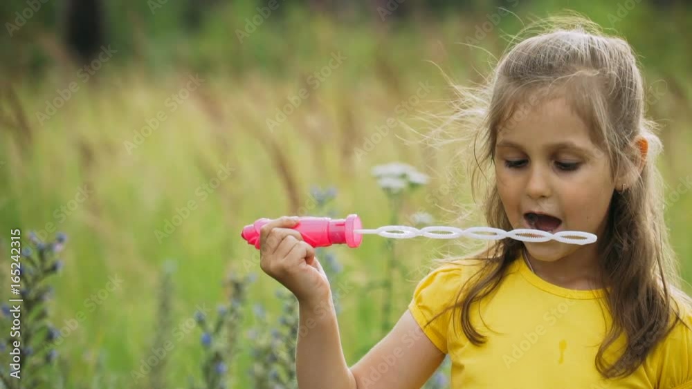 Cute little girl blowing soap bubbles on a summer meadow against the background of tall grass and flowers. Slow motion. Cute little girl blowing soap bubbles on a summer meadow against the background of tall grass and flowers. Slow motion.