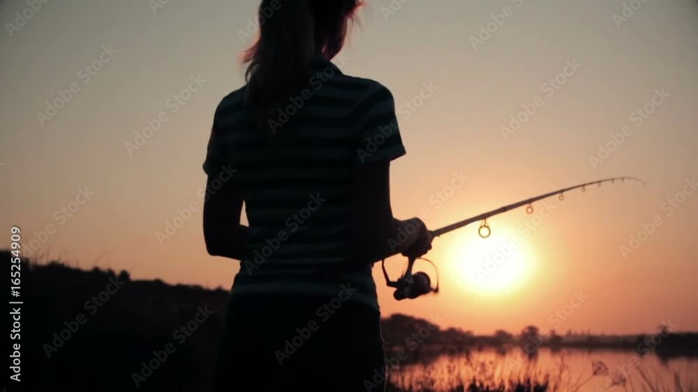 Silhouette of a young woman resting on nature fishing on a fishing rod by spinning at dawn