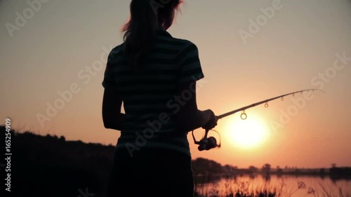 Silhouette of a young woman resting on nature fishing on a fishing rod by spinning at dawn