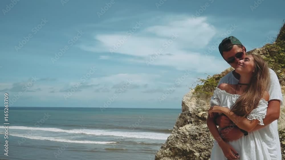 Romantic young couple hugging on shore cliff on a sunny summer day with blue sky, Bali, camera moving around them in slow motion. Shot with sony a7s on a motorized 3-axis handheld gimbal.