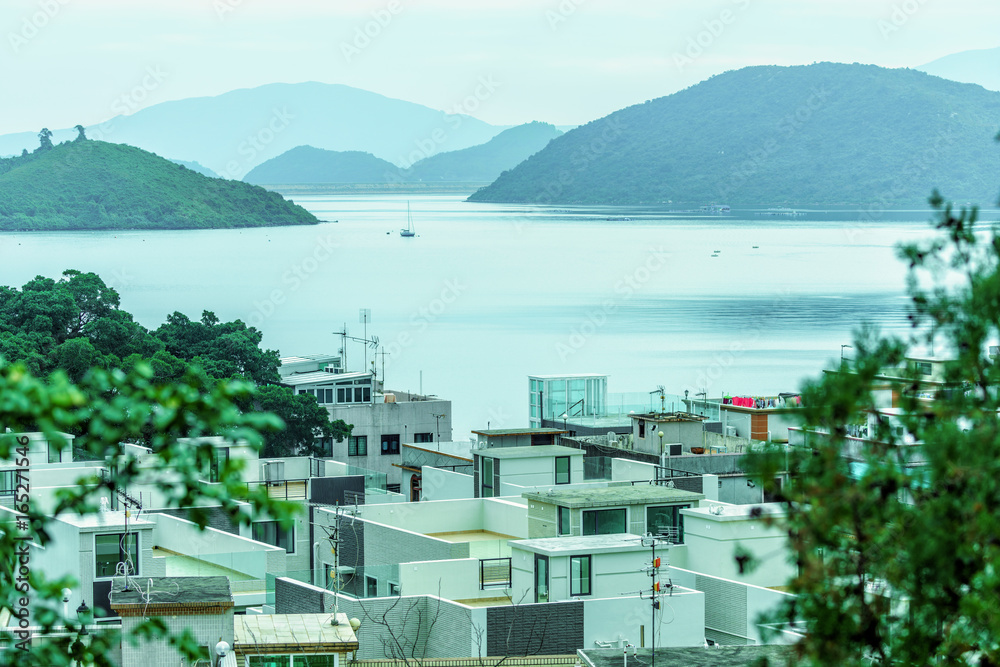 Obraz premium Hong Kong New Territories residential housing view with distant sailboat floating on lake water on green mountains background. Scenic landscape of Sha Lan villas and Plover Cove at Shuen Wan