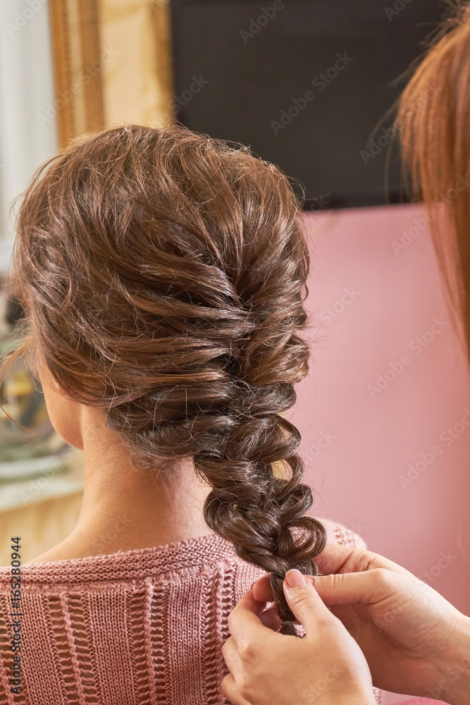 Hands of hairdresser plaiting braid. Female hairdo back view. Braid designs for women.