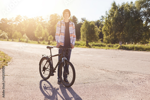 Fashionable hipster teenager wearing shirt and jeans standing on road with bicycle enjoying his hobby. Young cyclist having rest after riding bicycle. People, lifestyle and recreation concept