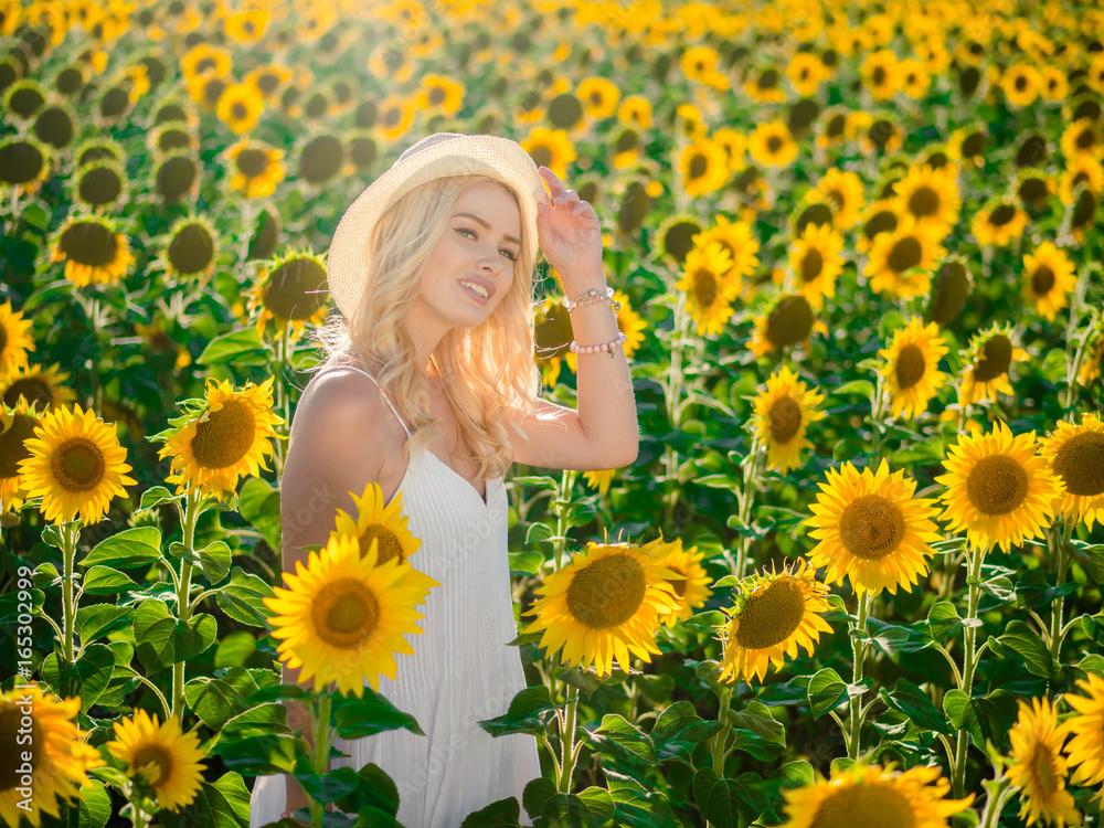 Young beautiful blonde woman standing in sunflower field. Sunset background. Sexy sensual portrait of girl in straw hat and white summer dress. Young beautiful blonde woman standing in sunflower field. Sunset background. Sexy sensual portrait of girl in straw hat and white summer dress.