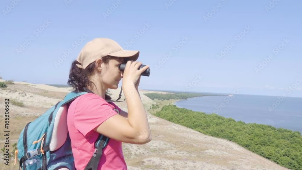 Travel woman looking at wildlife with binoculars. Woman on vacation looking at nature landscape. Hiker with a backpack is looking through binoculars at sea, at beautiful landscape. Mountain. Beach. Travel woman looking at wildlife with binoculars. Woman on vacation looking at nature landscape. Hiker with a backpack is looking through binoculars at sea, at beautiful landscape. Mountain. Beach.
