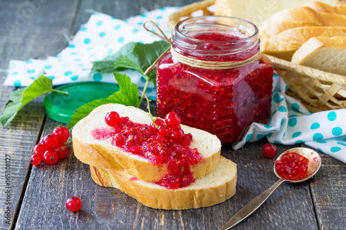 Homemade jam. A glass jar with red currant jam and white bread on a kitchen wooden table. Preserved berry.