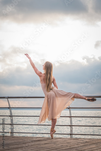 Dancing ballerina in beige silk dress and pointe on embankment above ocean or sea beach at sunrise. Young beautiful blonde woman with long hair practicing classic exercises with emotions.