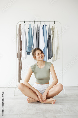 Portrait of cheerful young pretty brunette girl smiling looking at camera sitting on floor over hanger wardrobe and white wall background. photo