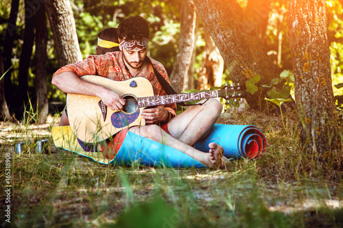 A young, unshaven male hippie outdoors sitting on the grass and plays the guitar behind his girlfriend sits and listens