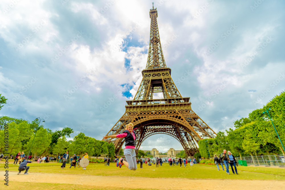 Tourist woman jumping at Tour Eiffel, icon of Paris. Caucasian lifestyle traveler enjoing at Eiffel Tower from Champ de Mars garden in Paris, France. Freedom and travel concept in European Capitals.
