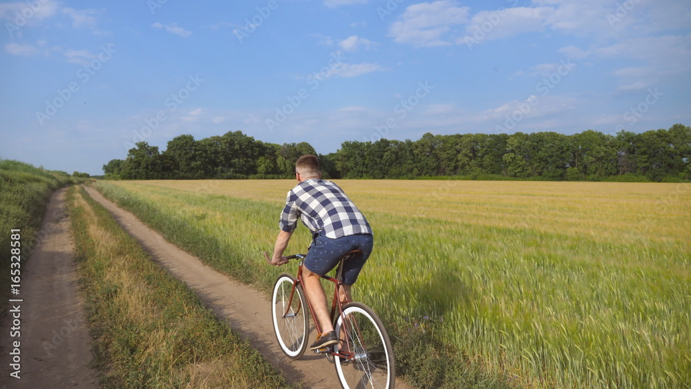 Young man riding vintage bicycle at the rural road over field. Sporty guy cycling along country trail outdoor. Male cyclist riding bike in the countryside. Healthy active lifestyle