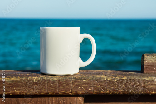 White mug, mock up, empty space for artwork, text, standing on wood plank, turquoise sea, clear blue sky, horizon, sunlight, nature