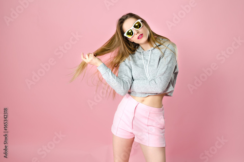 Portrait of young attractive funny teenager girl with shrtaight blonde hair and sunglasses in trendy clothes on the pink background, studio shooting. 