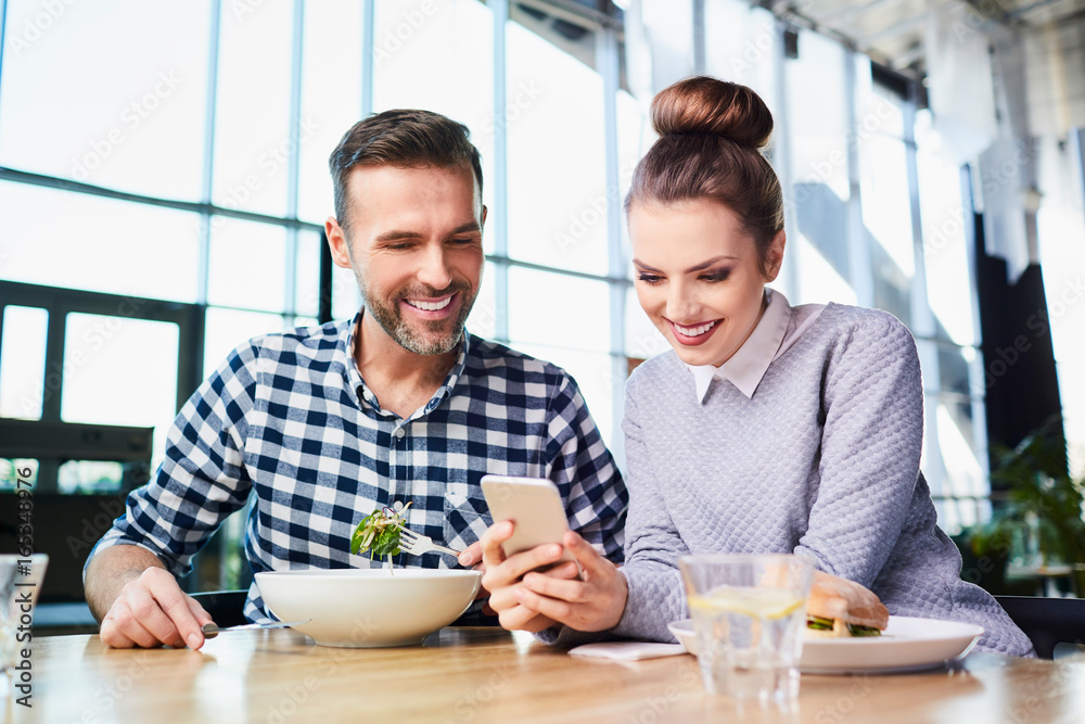 Happy middle-aged couple eating lunch together and looking at phone Happy middle-aged couple eating lunch together and looking at phone