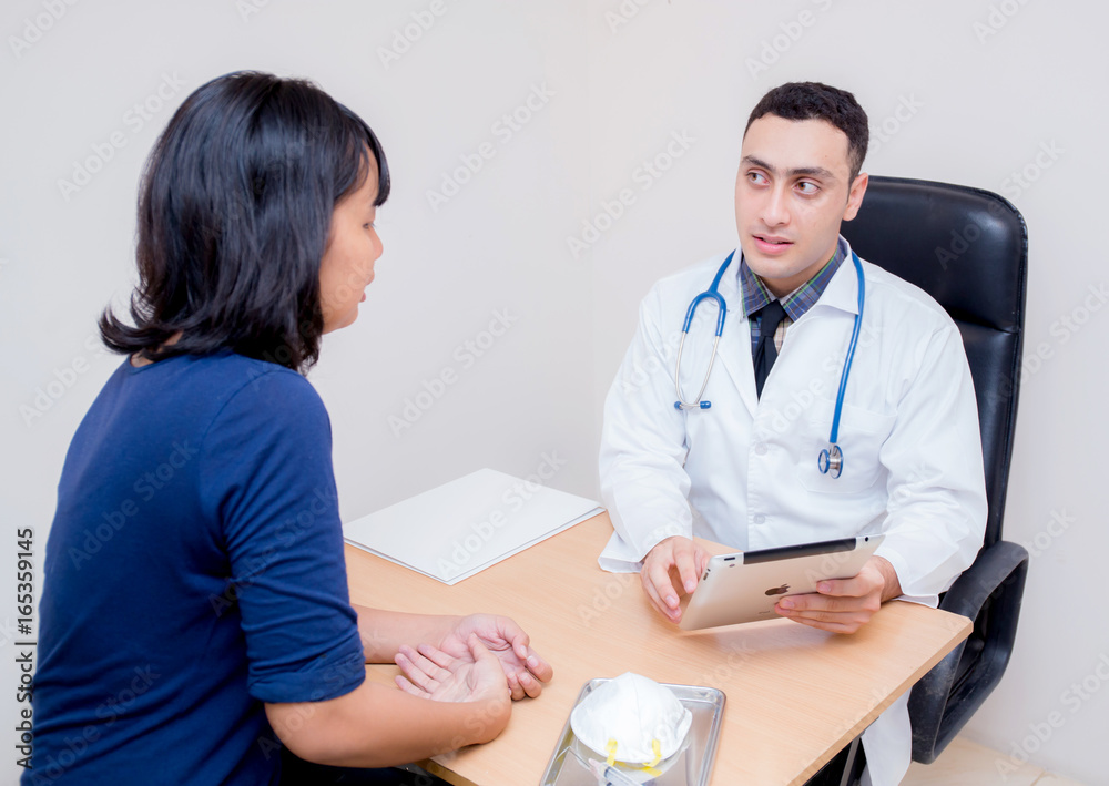 Asian man doctor holding tablet discussing with asian woman patient about examination result at table in clinic. Asian man doctor holding tablet discussing with asian woman patient about examination result at table in clinic.