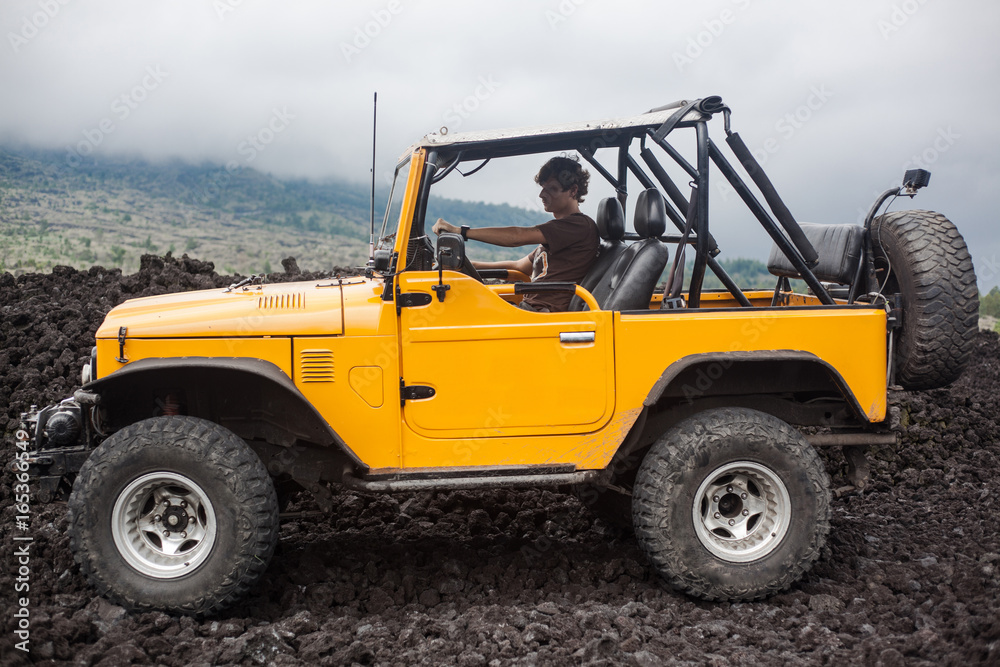 Φωτογραφία A curly-haired man is sitting in the offroad yelow vehicle turned to the right and parked at top of a valley with volcanic rock and mountains