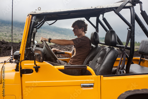 A curly-haired man is sitting in the offroad yelow vehicle turned to the right and parked at top of a valley with volcanic rock and mountains in Bali, Indonesia