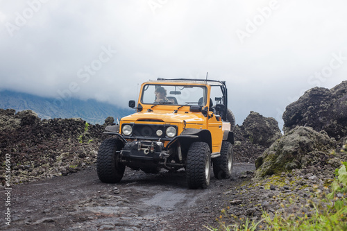 A curly-haired man is looking away driving an offroad yelow vehicle at the top of a valley with dark ground road photo
