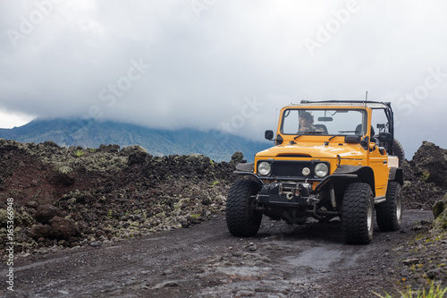 A curly-haired man is looking away driving an offroad yelow vehicle at the top of a valley with dark ground road